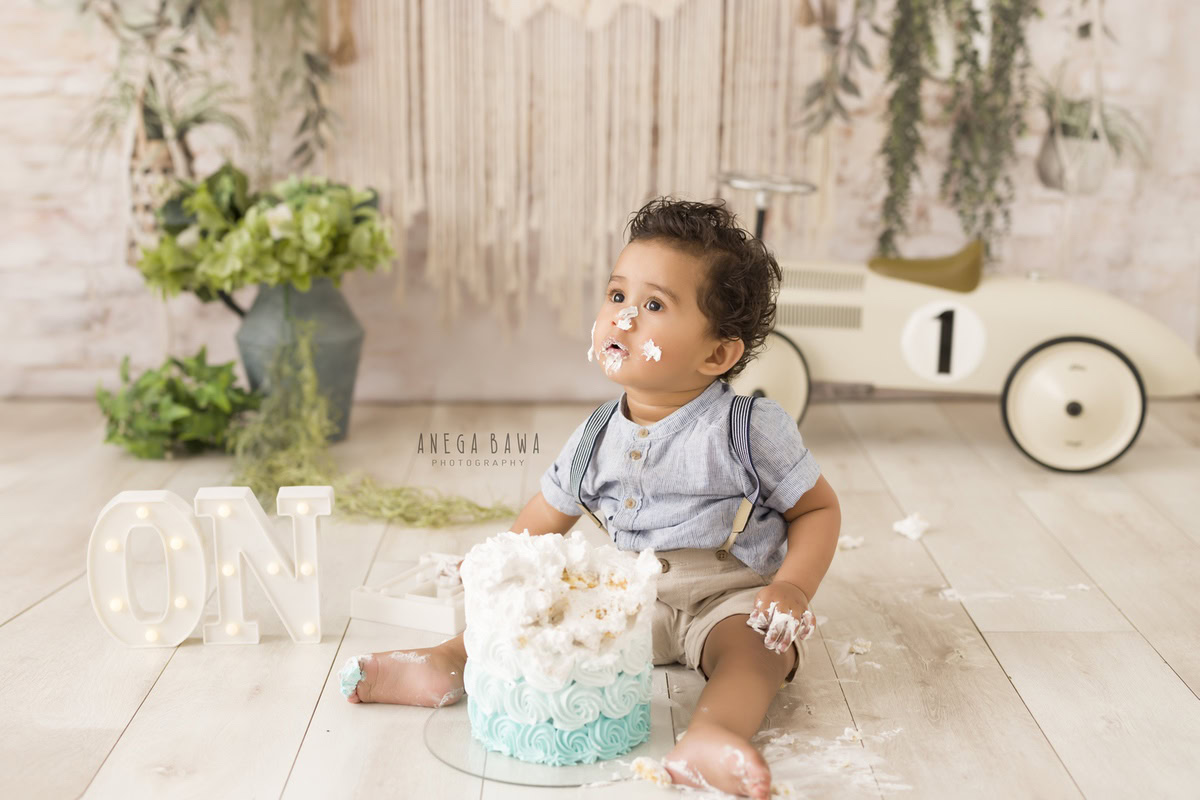 1year old boy with a toy scooter and cake smash setup, featuring a green flower vase, against a beige backdrop, captured by Anega Bawa during a Cake Smash photoshoot in Delhi, Gurgaon
