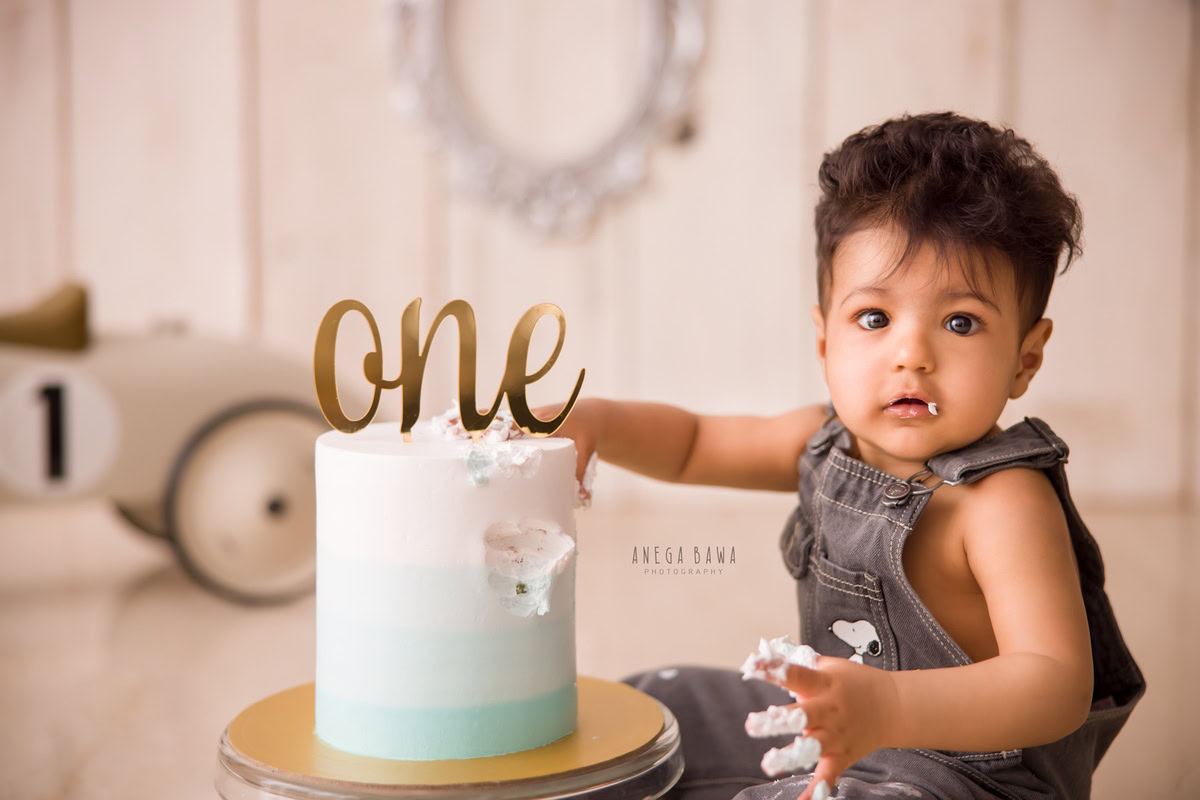 1 year old boy in denim dungarees with a toy scooter, enjoying a cake smash setup featuring a silver frame on the wall. Celebrating his first birthday. Cake Smash photo shoot in Delhi, Gurgaon by Anega Bawa Family Photographer Gurgaon (Delhi NCR).