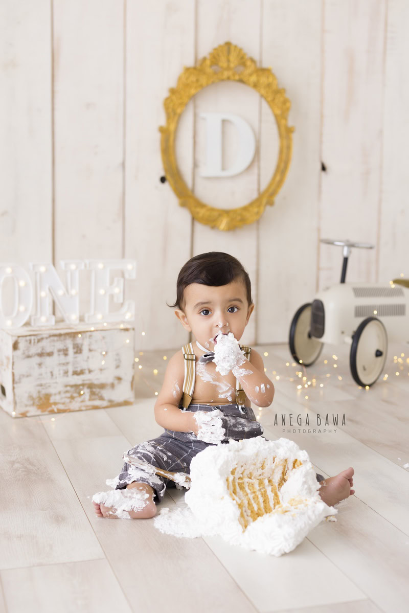 1year old boy with a toy scooter and cake smash setup, featuring a golden alphabet frame on the wall, against a beige backdrop, captured by Anega Bawa during a Cake Smash photoshoot in Delhi, Gurgaon