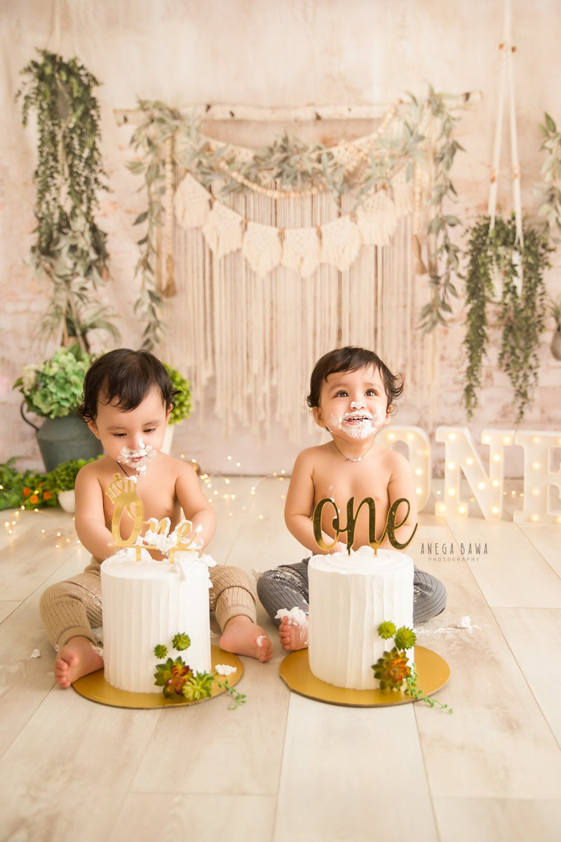 1 year old boy with twin Cake Smash setups, beige and white backdrop, green vase, leafy fringe, first birthday photo shoot in Delhi, Gurgaon. Captured by Anega Bawa family photographer Gurgaon (Delhi NCR)