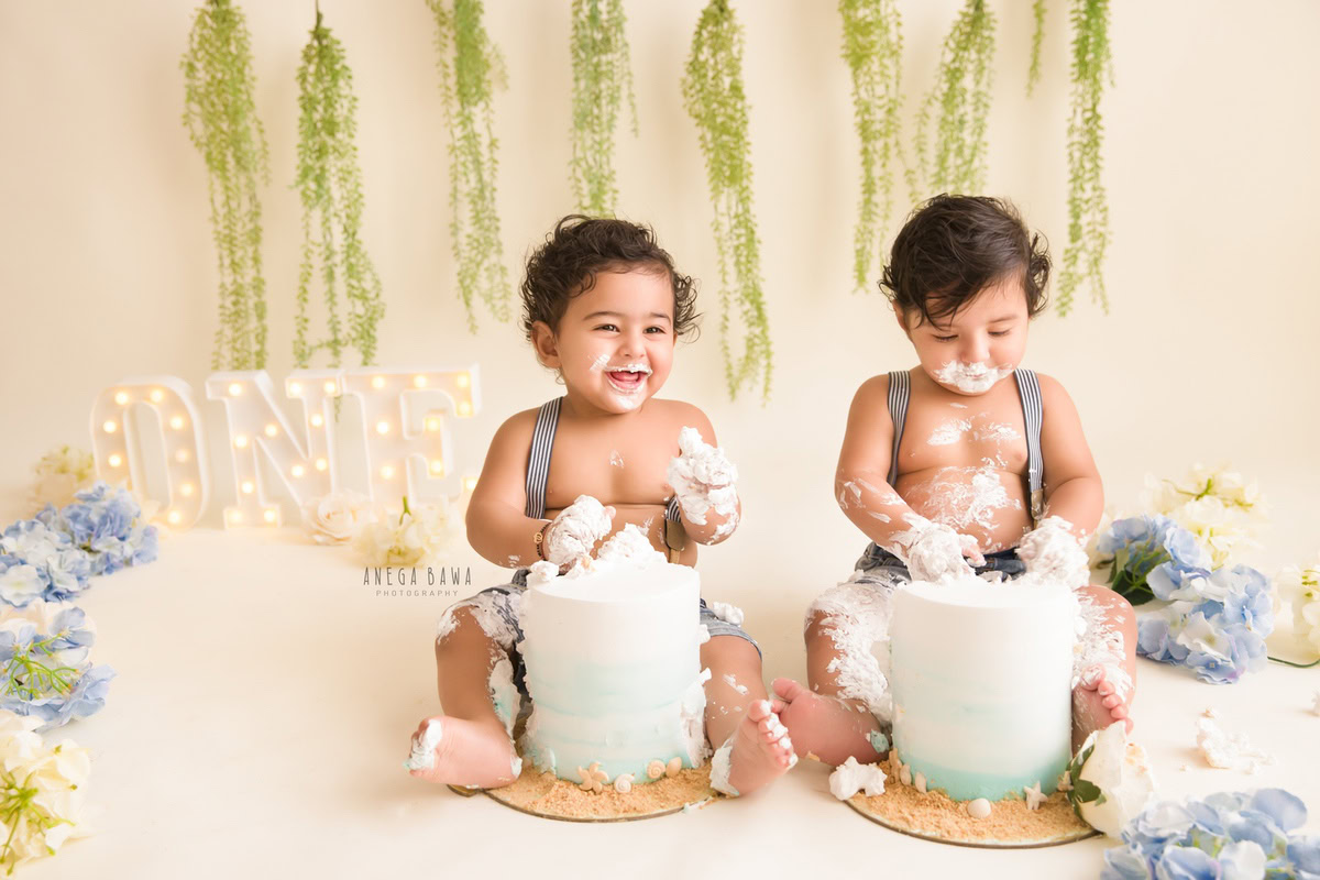 1 year old boy twin in denim dungaree, surrounded by flowers on the floor and a leafy fringe on the wall, playing with cake smash. Captured during a delightful first birthday photography session in Delhi Gurgaon by the renowned family photographer Anega Bawa.