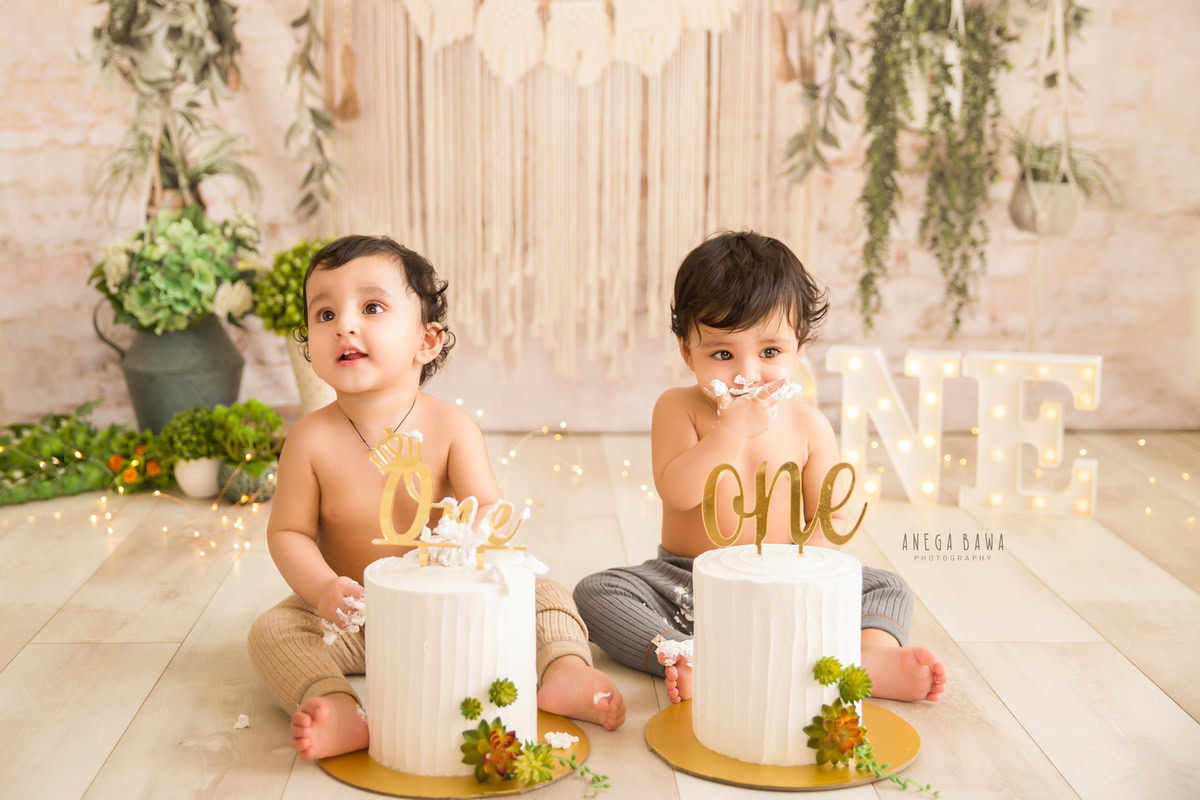1 year old twins with two cake smash setups, featuring a beige backdrop, leafy fringe, and fairy lights. Celebrating their first birthday. Cake Smash photo shoot in Delhi, Gurgaon by Anega Bawa Family Photographer Gurgaon (Delhi NCR).