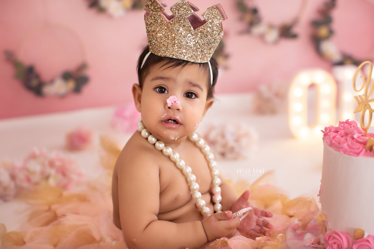1 year old boy wearing a crown with pearls, pink backdrop, cake smash, and flowers on the floor. Captured during a charming first birthday photoshoot in Delhi Gurgaon by the renowned family photographer Anega Bawa.