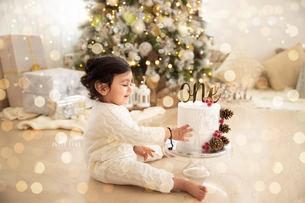 1 year old boy in a white babysuit with a Christmas tree, fairy lights, and a white backdrop, enjoying a cake smash setup. Celebrating his first birthday. Cake Smash photo shoot in Delhi, Gurgaon by Anega Bawa Family Photographer Gurgaon (Delhi NCR).