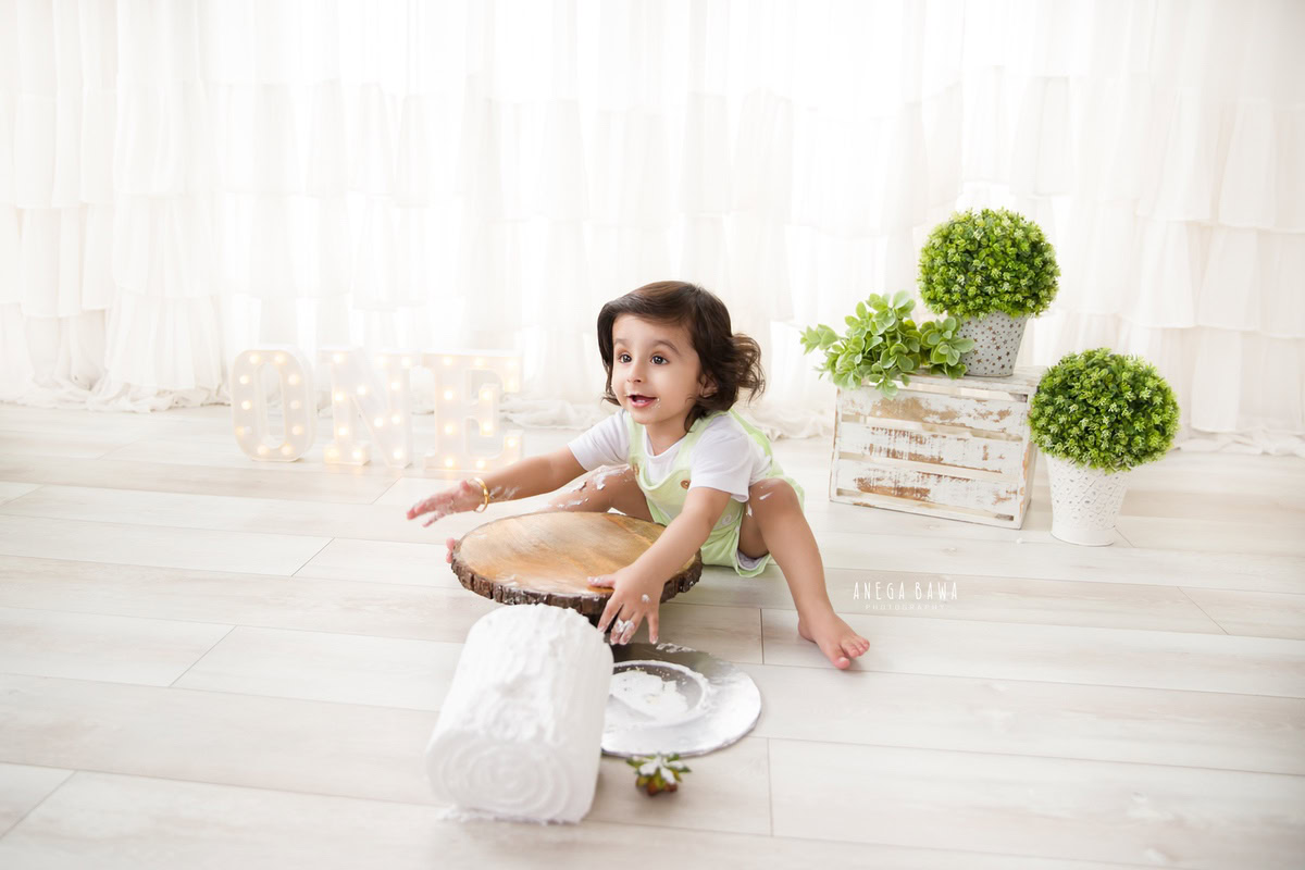 1 year old boy with cake smash, white backdrop, pots with leaves. Captured during a delightful first birthday photoshoot in Delhi Gurgaon by the renowned family photographer Anega Bawa.