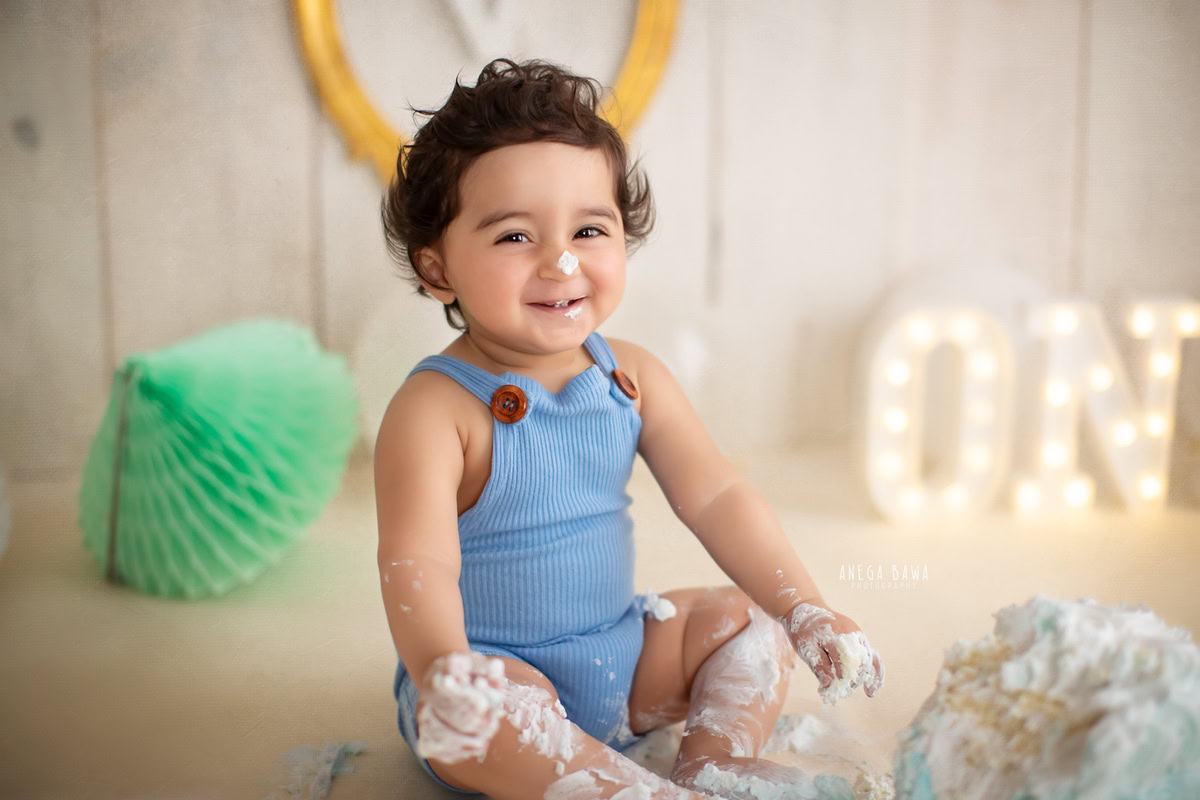 1 year old boy with cake smash, beige backdrop, and pompom decorations. Captured during a charming first birthday photoshoot in Delhi Gurgaon by the well-known family photographer Anega Bawa.