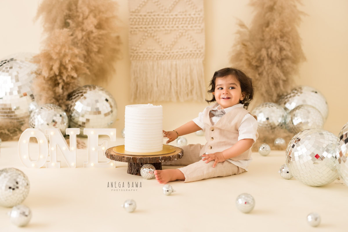 Adorable toddler celebrating first birthday with cake smash, surrounded by festive decorations, silver balloons, and a white cake, capturing joyful moments for memorable photography.