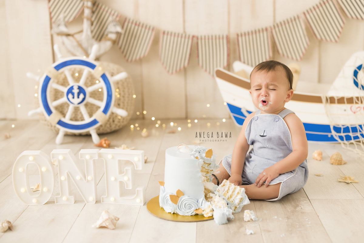 1 year old boy with cake smash in cute pout pose with beige backdrop and fairy lights with toy spaceship. Captured during a fun first birthday photography session in Delhi Gurgaon by the esteemed family photographer Anega Bawa.