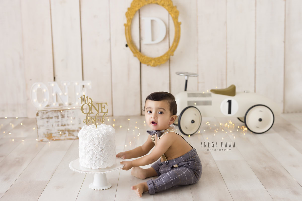 1 year old boy with a cake smash setup, featuring a toy scooter, a beige backdrop, and fairy lights. Celebrating his first birthday. Cake Smash photo shoot in Delhi, Gurgaon by Anega Bawa Family Photographer Gurgaon (Delhi NCR).