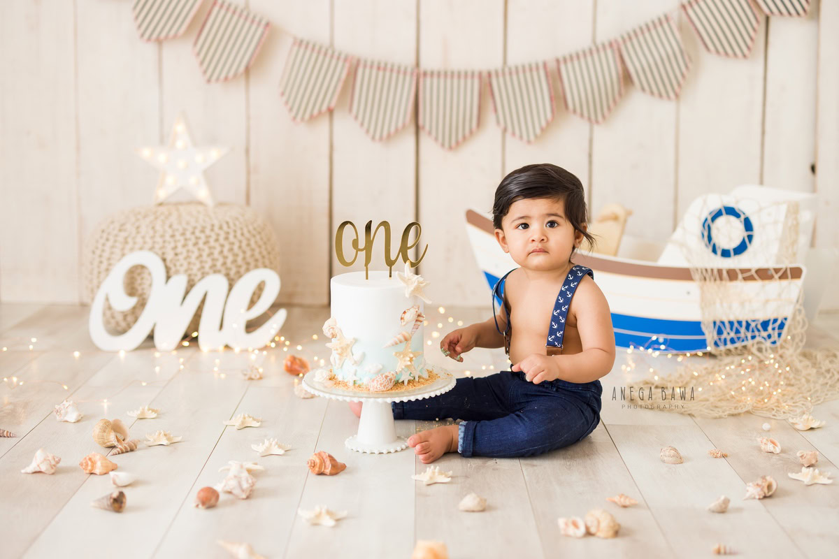 1 year old boy with a cake smash setup, featuring a toy spaceship, flowers on the floor, and a beige backdrop. Celebrating his first birthday. Cake Smash photo shoot in Delhi, Gurgaon by Anega Bawa Family Photographer Gurgaon (Delhi NCR).