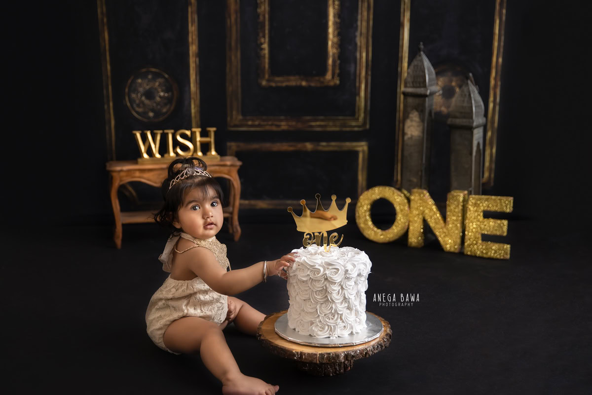 1 year old girl with a cake smash setup, featuring a brown backdrop, a golden alphabet frame on the floor, and a castle. Celebrating her first birthday. Cake Smash photo shoot in Delhi, Gurgaon by Anega Bawa Family Photographer Gurgaon (Delhi NCR).