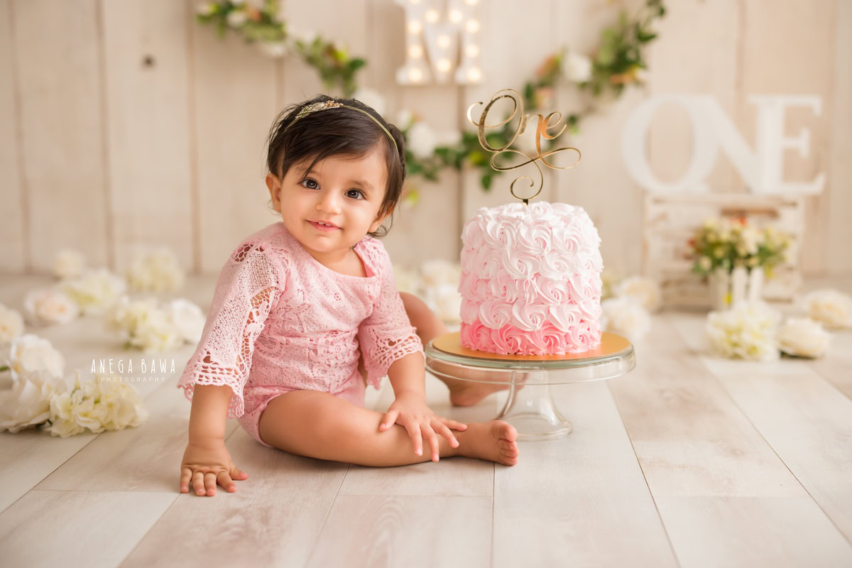1 year old girl with a cake smash setup, featuring an alphabet light frame on the wall, floral fringe, white flowers, and a beige backdrop. Celebrating her first birthday. Cake Smash photo shoot in Delhi, Gurgaon by Anega Bawa Family Photographer Gurgaon (Delhi NCR).