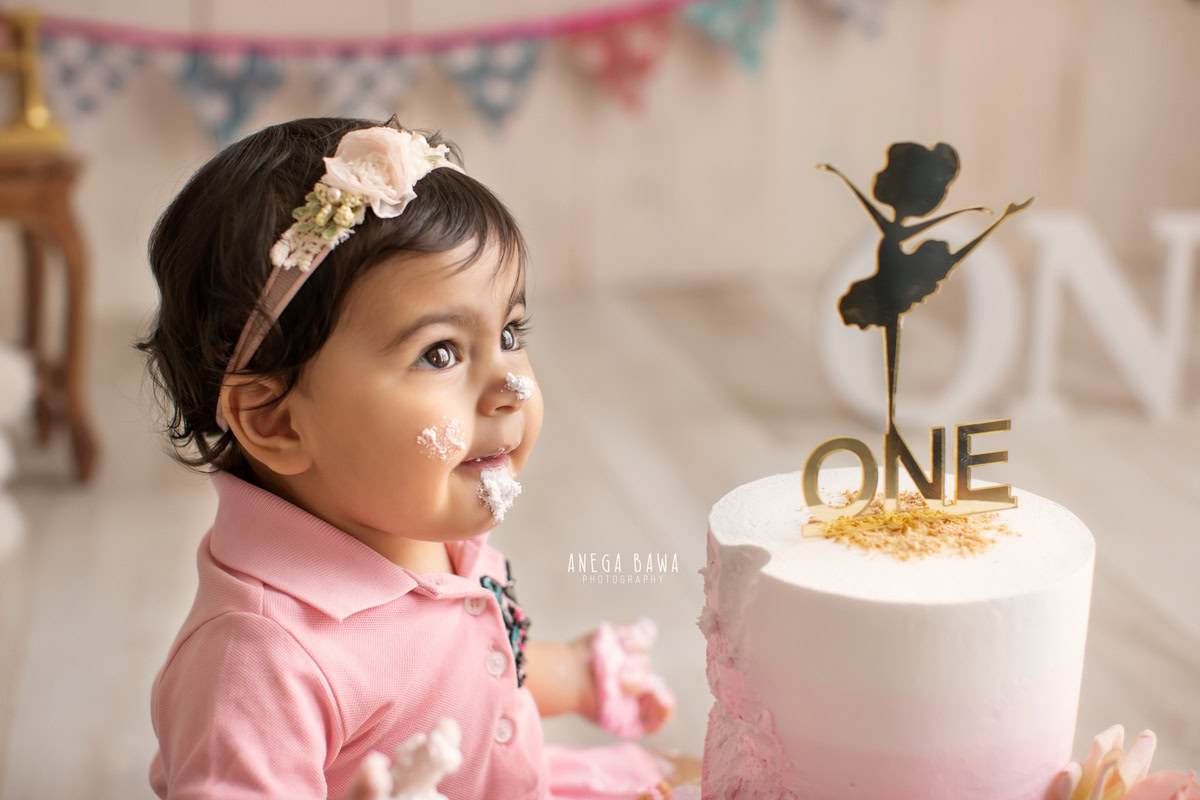 1 year old girl with cake smash, beige backdrop, and cute headband. Captured during a delightful first birthday photography session in Delhi Gurgaon by the acclaimed family photographer Anega Bawa.