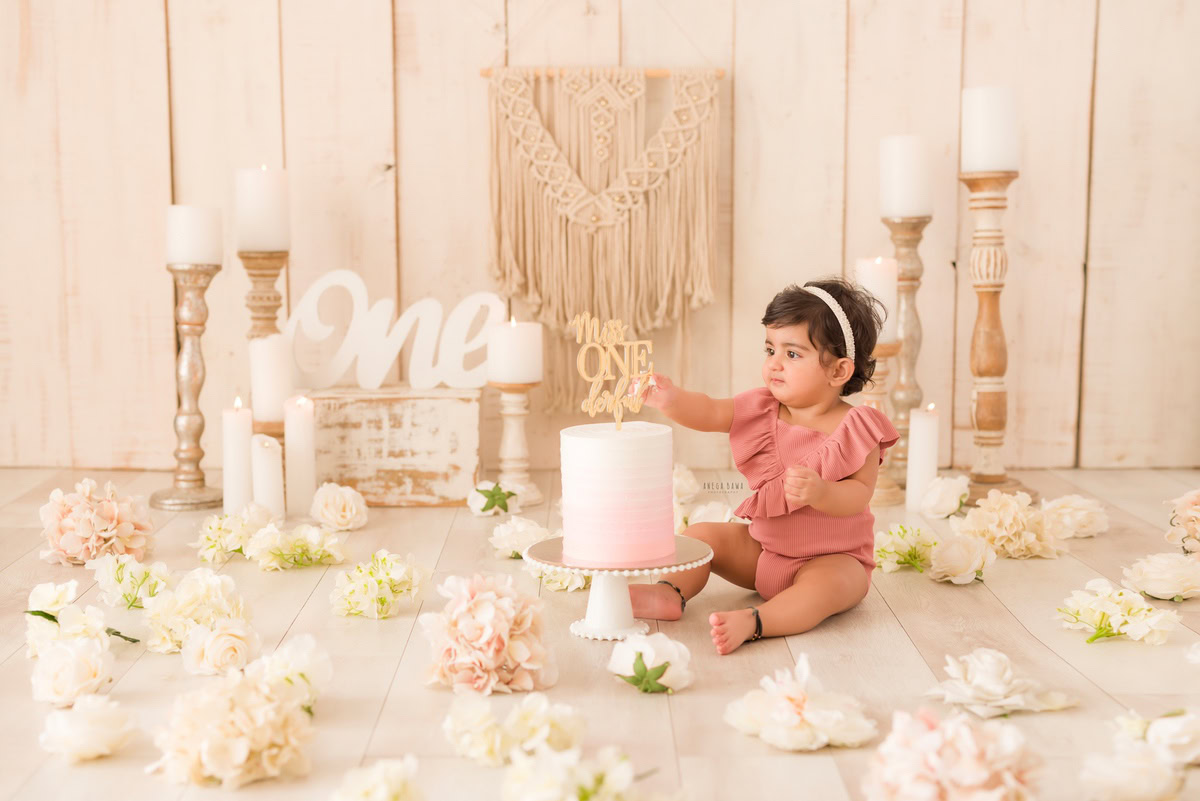 1 year old girl Cake Smash featuring a charming candle stand, delicate ivory flowers on the floor, and a soft beige backdrop, first birthday photo shoot in Delhi, Gurgaon. Captured by Anega Bawa family photographer Gurgaon (Delhi NCR)