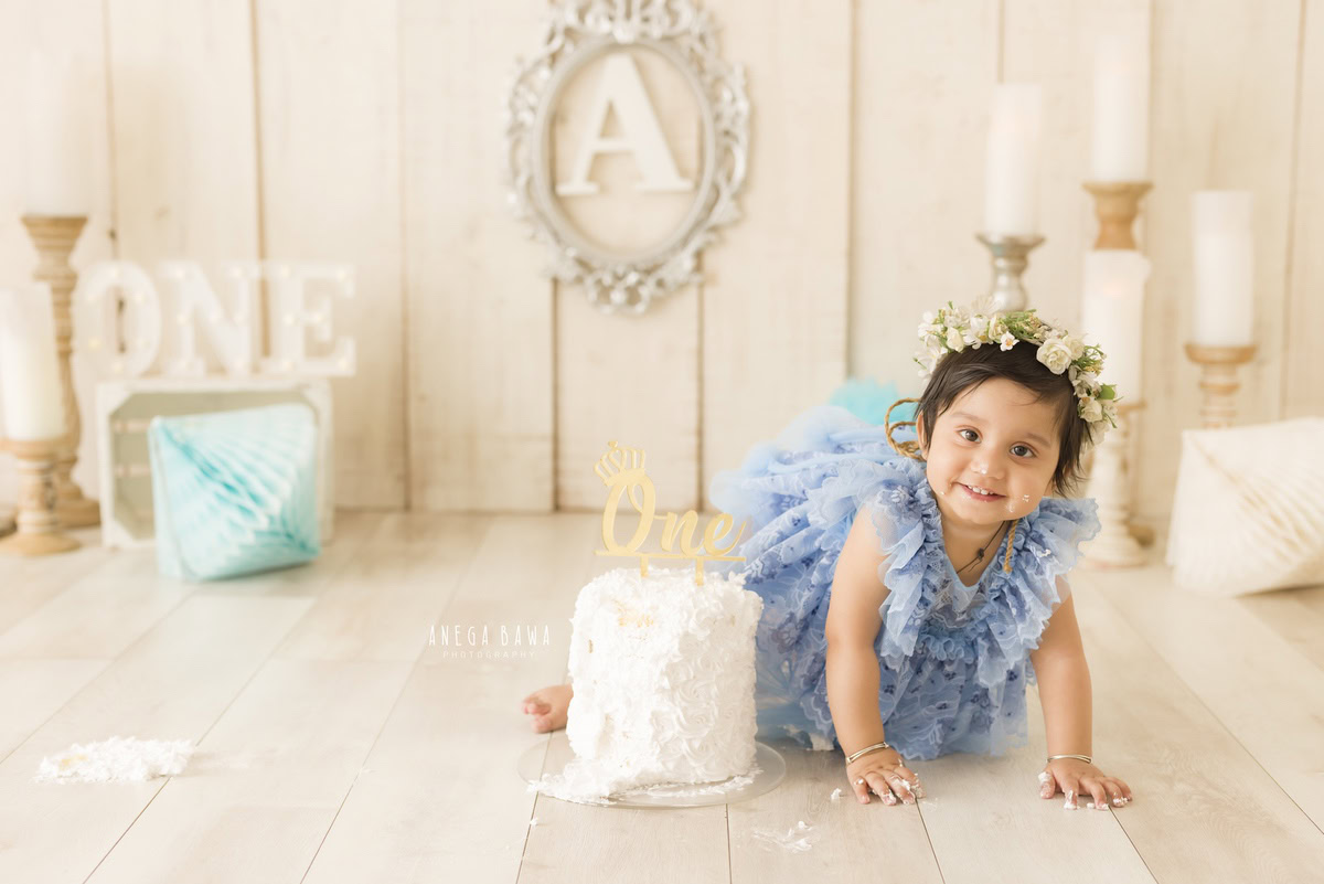 1year old girl with a cake smash setup, featuring a candle stand, pom-pom decorations, and a silver alphabet frame on the wall, captured by Anega Bawa during a Cake Smash photoshoot in Delhi, Gurgaon