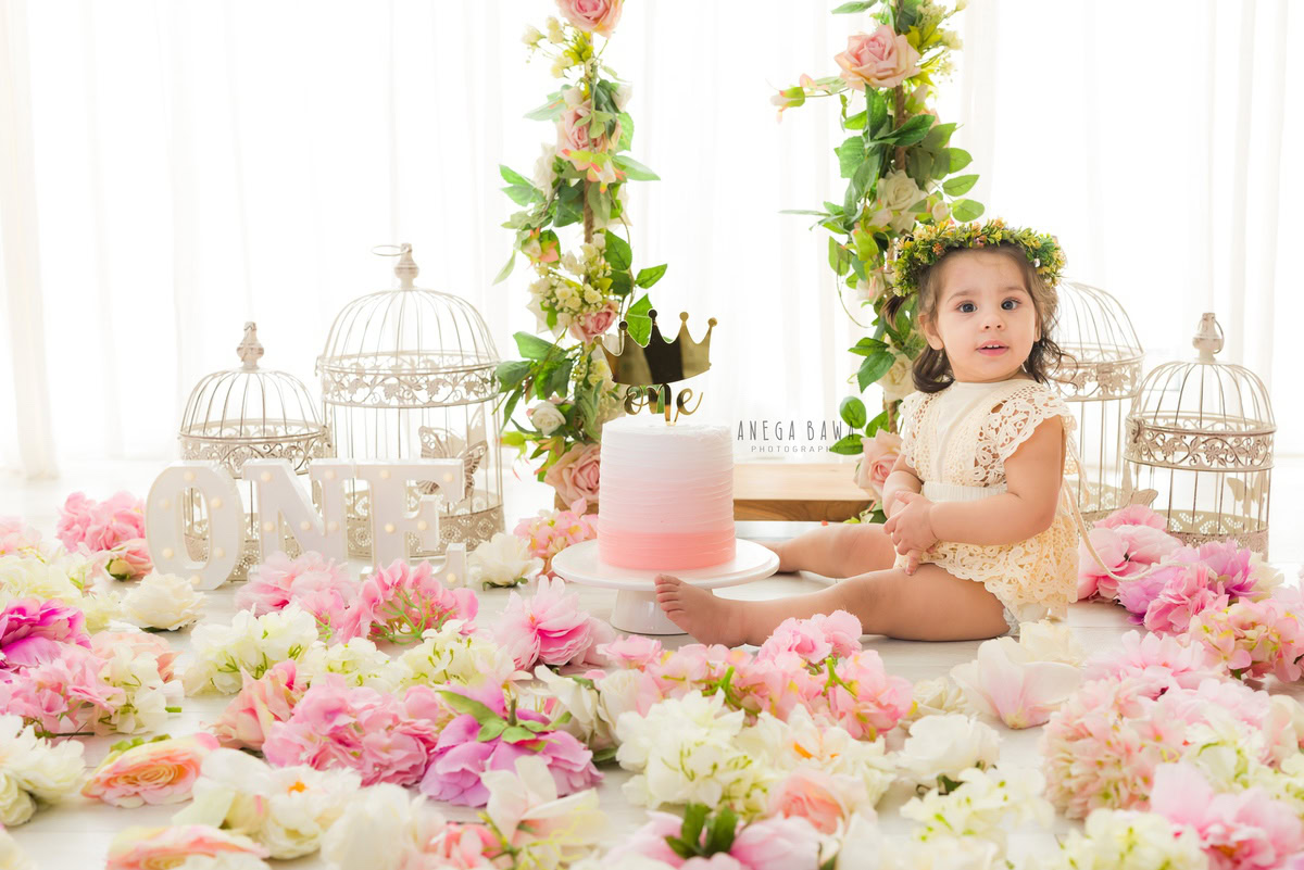 1year old girl with a cake smash setup featuring castles and pink and white flowers on the floor against a white backdrop, captured by Anega Bawa during a Cake Smash photoshoot in Delhi, Gurgaon