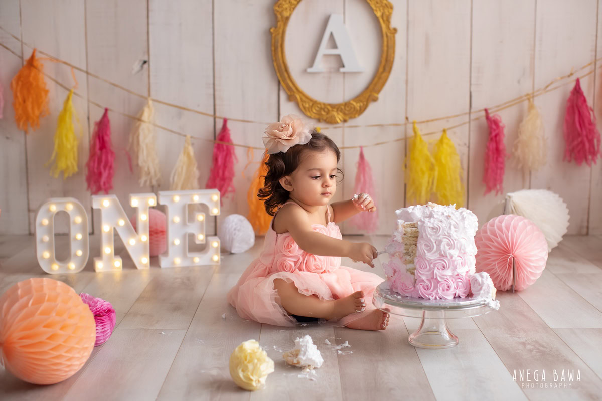 1 year old girl with cake smash, colorful fringe on the wall, pompom decorations, and beige backdrop. Captured during a vibrant first birthday photo shoot in Delhi Gurgaon by the esteemed family photographer Anega Bawa.