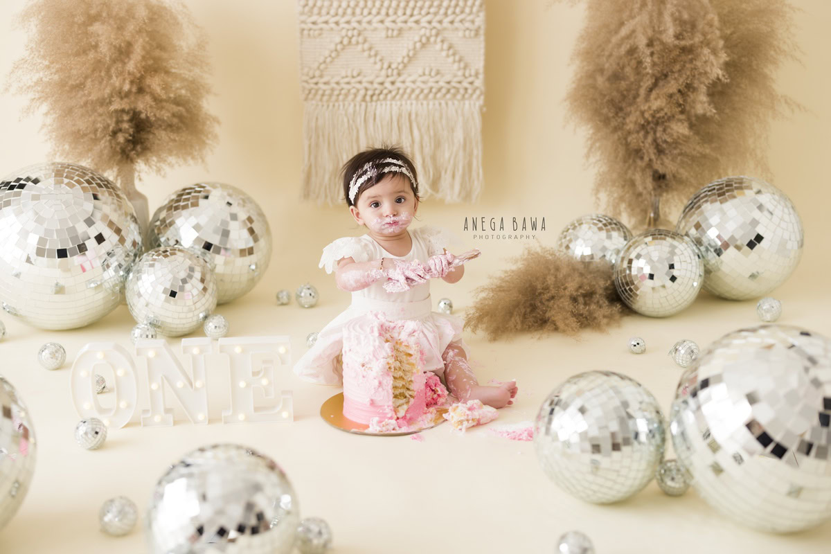 1 year old girl with cake smash, discoballs, and bushes against a beige backdrop. Captured during a lively first birthday photography session in Delhi Gurgaon by the renowned family photographer Anega Bawa.