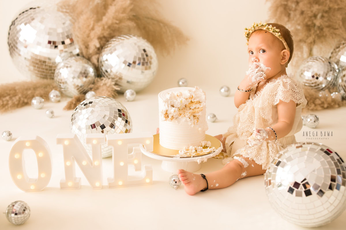 1 year old girl with a cake smash setup, featuring disco balls and bushes against a beige backdrop. Celebrating her first birthday. Cake Smash photo shoot in Delhi, Gurgaon by Anega Bawa Family Photographer Gurgaon (Delhi NCR).