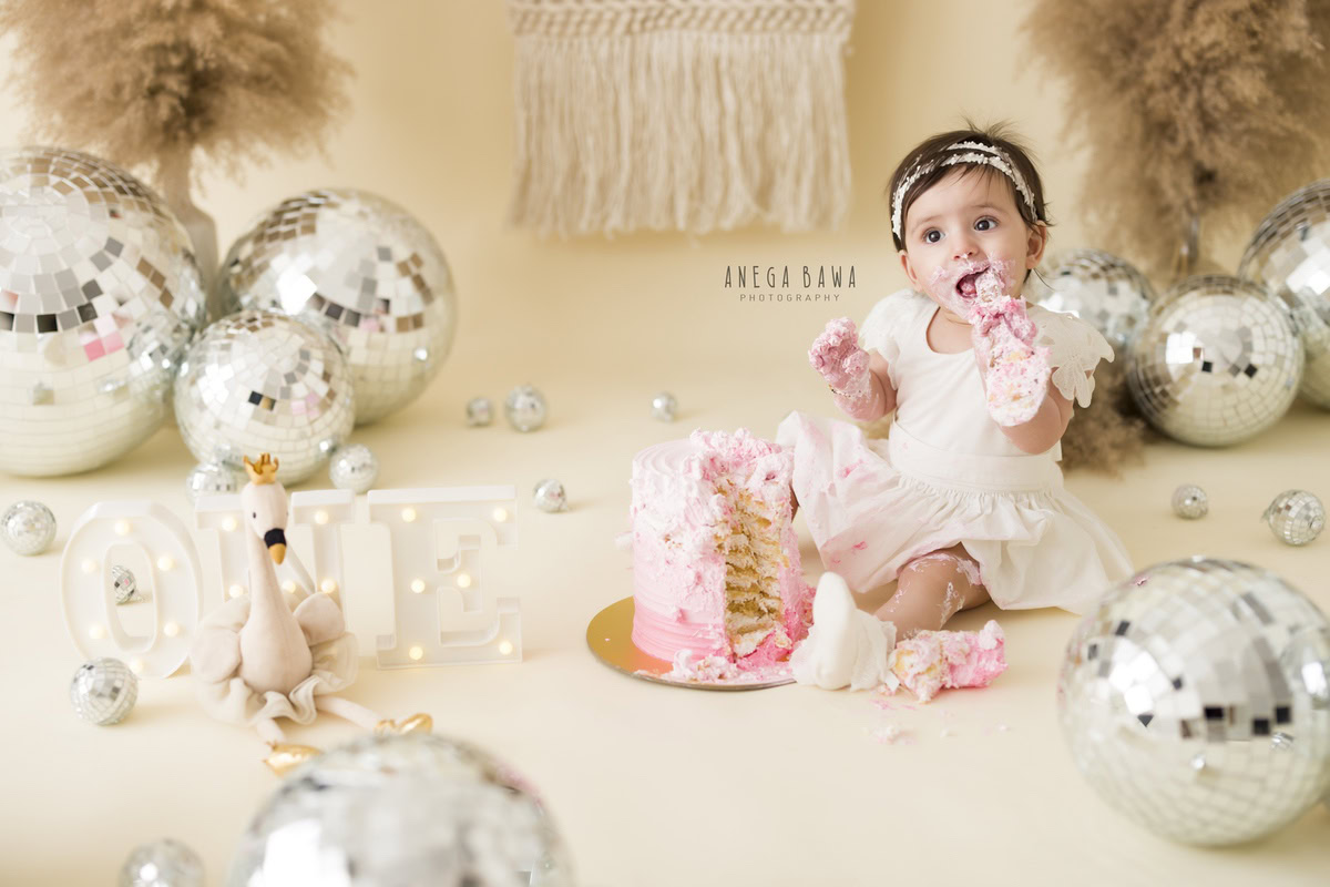 1 year old girl with a cake smash setup, featuring disco balls and bushes against a beige backdrop. Celebrating her first birthday. Cake Smash photo shoot in Delhi, Gurgaon by Anega Bawa Family Photographer Gurgaon (Delhi NCR).