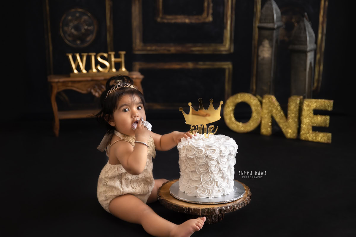 1 year old girl Cake Smash with a regal golden crown, rich brown backdrop, first birthday photo shoot in Delhi, Gurgaon. Captured by Anega Bawa family photographer Gurgaon (Delhi NCR)