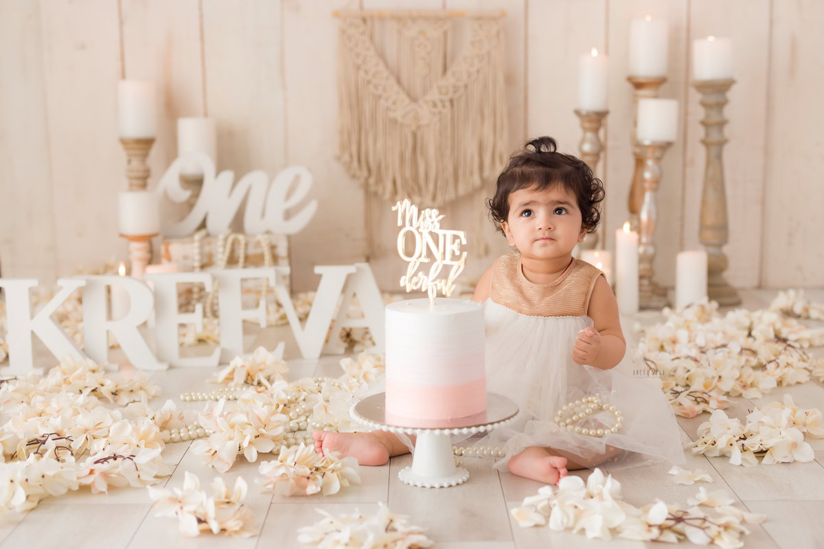 1year old girl with a cake smash setup featuring a name frame on the floor and a candle stand against a beige backdrop, captured by Anega Bawa during a Cake Smash photoshoot in Delhi, Gurgaon