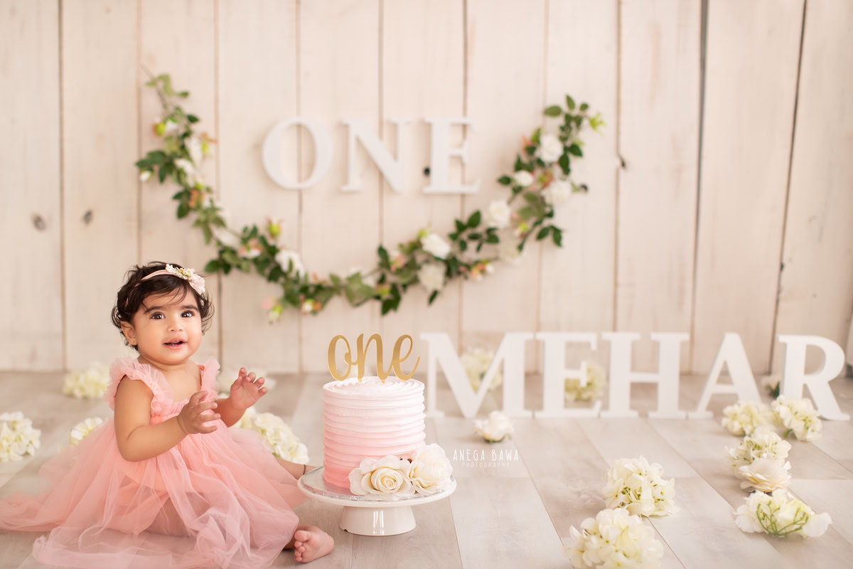 1 year old girl with a cake smash setup, featuring a name frame on the floor, leafy fringe on the wall, and a beige backdrop. Celebrating her first birthday. Cake Smash photo shoot in Delhi, Gurgaon by Anega Bawa Family Photographer Gurgaon (Delhi NCR).