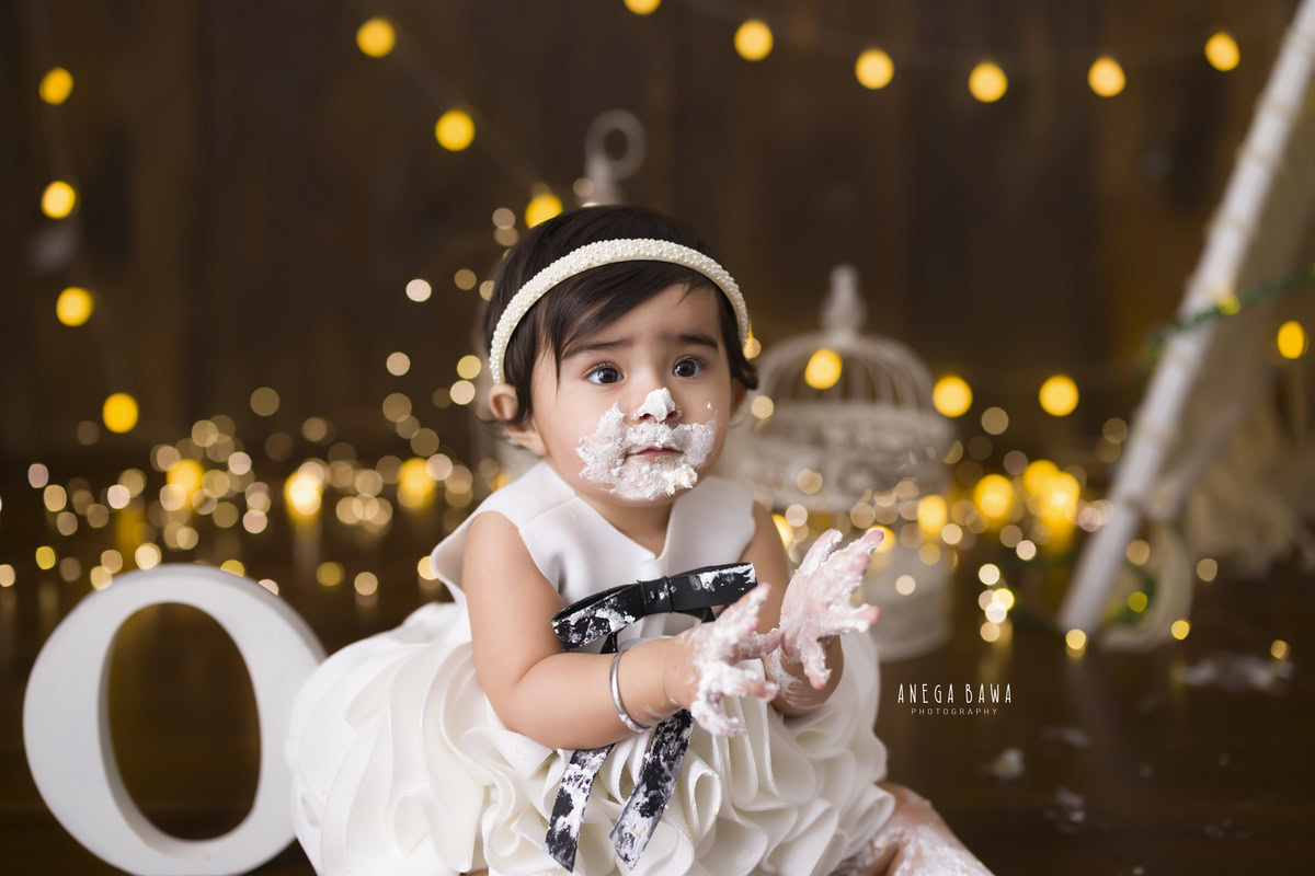 1 year old girl with cake smash on her face against a brown backdrop with fairy lights and a castle. Captured during a magical first birthday photoshoot in Delhi Gurgaon by the renowned family photographer Anega Bawa.