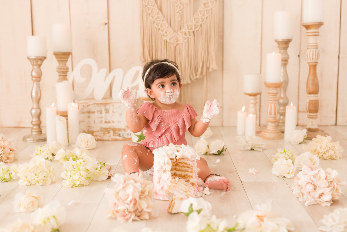 1year old girl with cake on her face, surrounded by a candle stand and flowers on the floor, against a beige backdrop, captured by Anega Bawa during a Cake Smash photoshoot in Delhi, Gurgaon