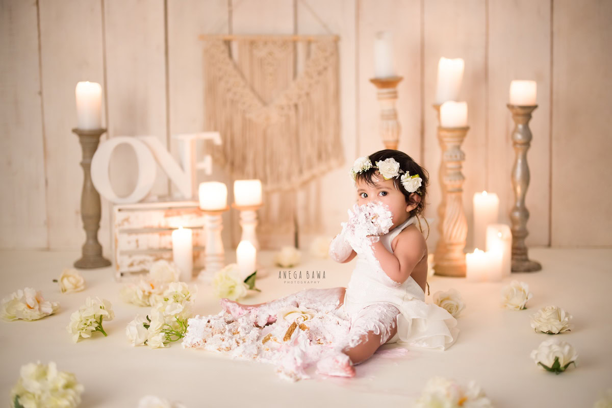 1 year old girl Cake Smash on face, with a charming candle stand, white candles, beige backdrop, and elegant white flowers, first birthday photography in Delhi, Gurgaon. Captured by Anega Bawa family photographer Gurgaon (Delhi NCR)