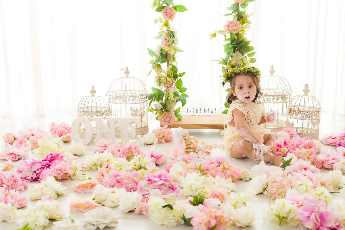 1 year old girl Cake Smash on face, surrounded by flowers on the floor, whimsical white castles, crisp white backdrop, and leafy fringes, first birthday photoshoot in Delhi, Gurgaon. Captured by Anega Bawa family photographer Gurgaon (Delhi NCR)