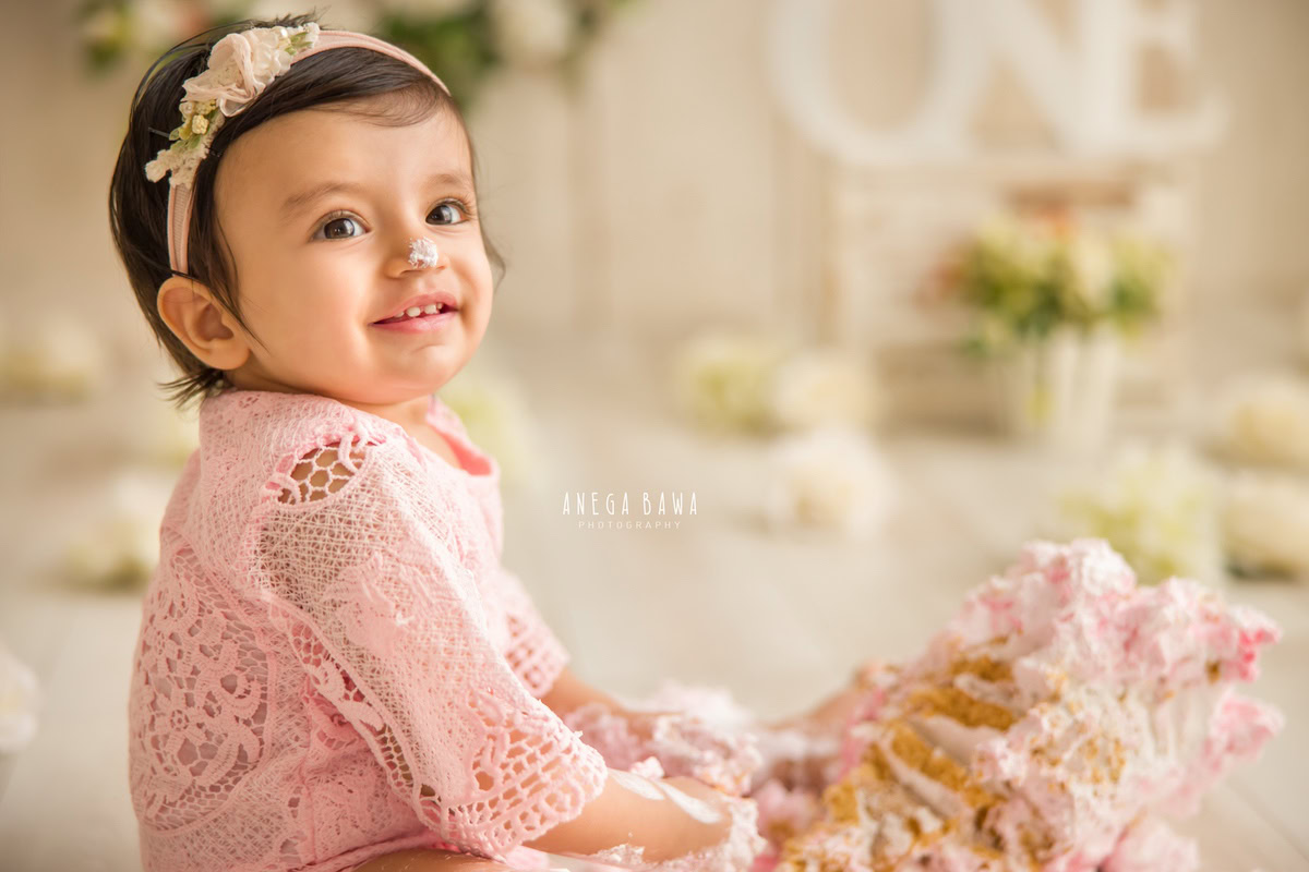 1 year old girl Cake Smash on nose, striking a cute pose, with a soft beige backdrop and flowers scattered on the floor, first birthday photography in Delhi, Gurgaon. Captured by Anega Bawa family photographer Gurgaon (Delhi NCR)
