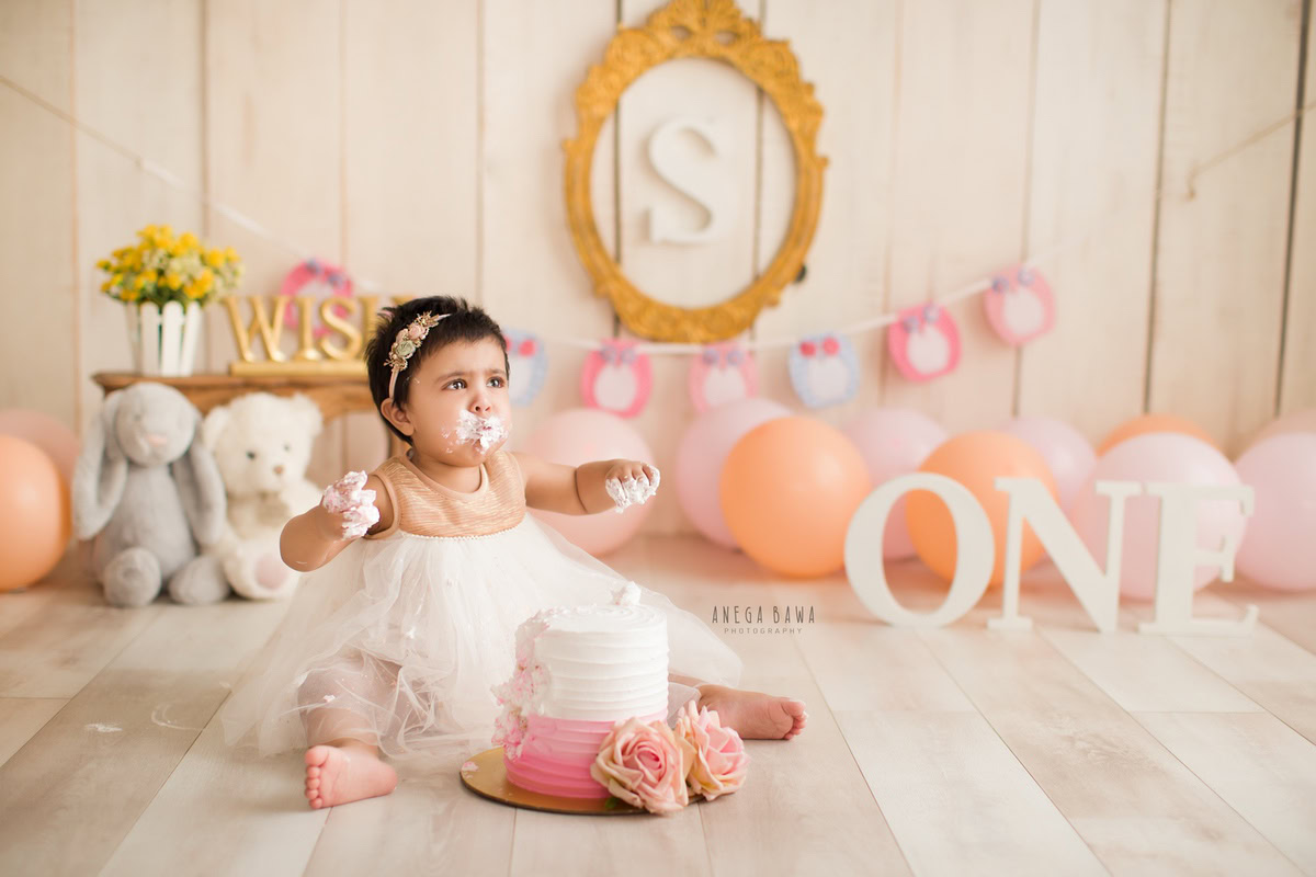 1 year old girl with a cake smash setup, featuring peach and pink balloons, teddy bears, and a beige backdrop. Celebrating her first birthday. Cake Smash photo shoot in Delhi, Gurgaon by Anega Bawa Family Photographer Gurgaon (Delhi NCR).