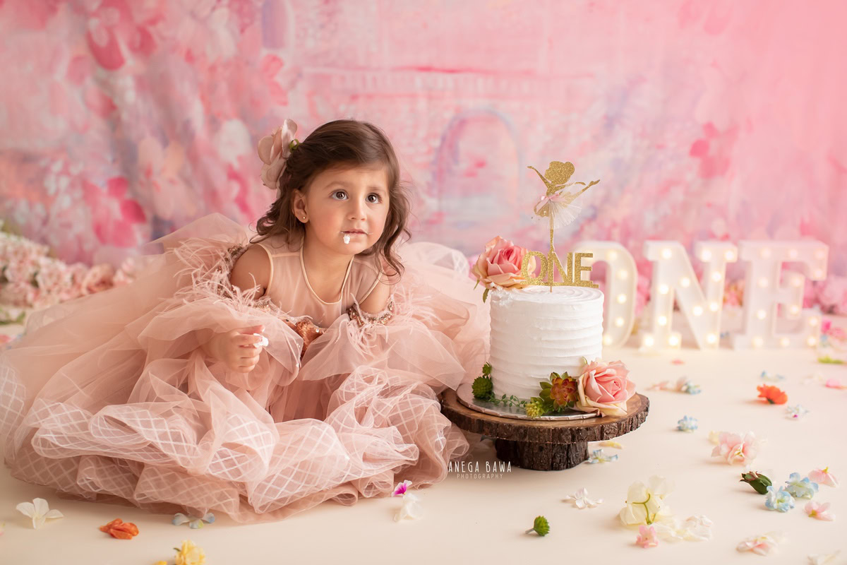 1 year old girl with cake smash against a pink backdrop with flowers on the floor. Captured during a lovely first birthday photoshoot in Delhi Gurgaon by the renowned family photographer Anega Bawa.