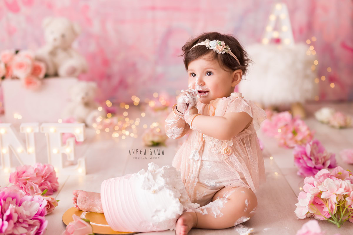 1 year old girl with cake smash against a pink backdrop with a white teddy bear, fairy lights, and pink and peach flowers on the floor. Captured during a charming first birthday photography session in Delhi Gurgaon by the renowned family photographer Anega Bawa.