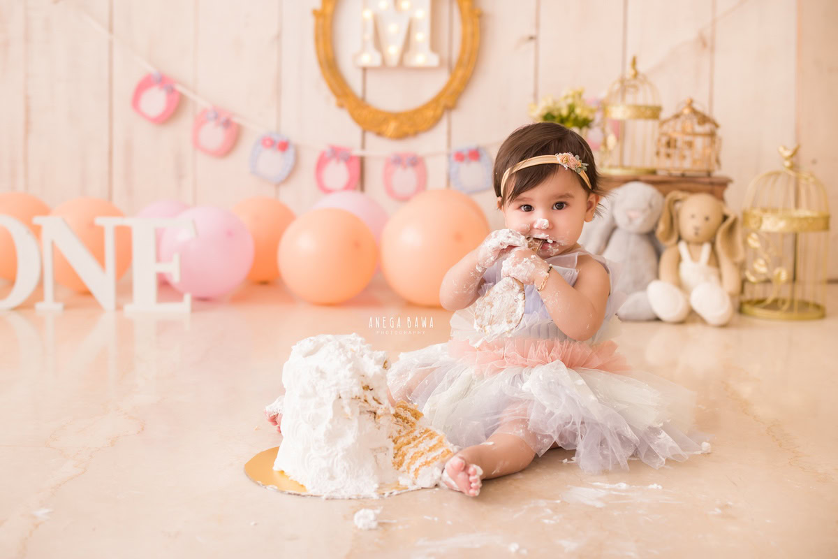 1 year old girl with cake smash surrounded by pink and peach balloons a golden castle teddybears and a golden alphabet frame on the wall. Captured during a delightful first birthday photoshoot in Delhi Gurgaon by the renowned family photographer Anega Bawa.