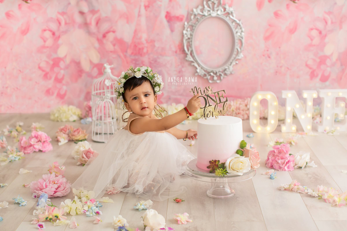 1 year old girl with a cake smash setup, featuring a silver frame on the wall, a pink backdrop, castles, and flowers on the floor. Celebrating her first birthday. Cake Smash photo shoot in Delhi, Gurgaon by Anega Bawa Family Photographer Gurgaon (Delhi NCR).