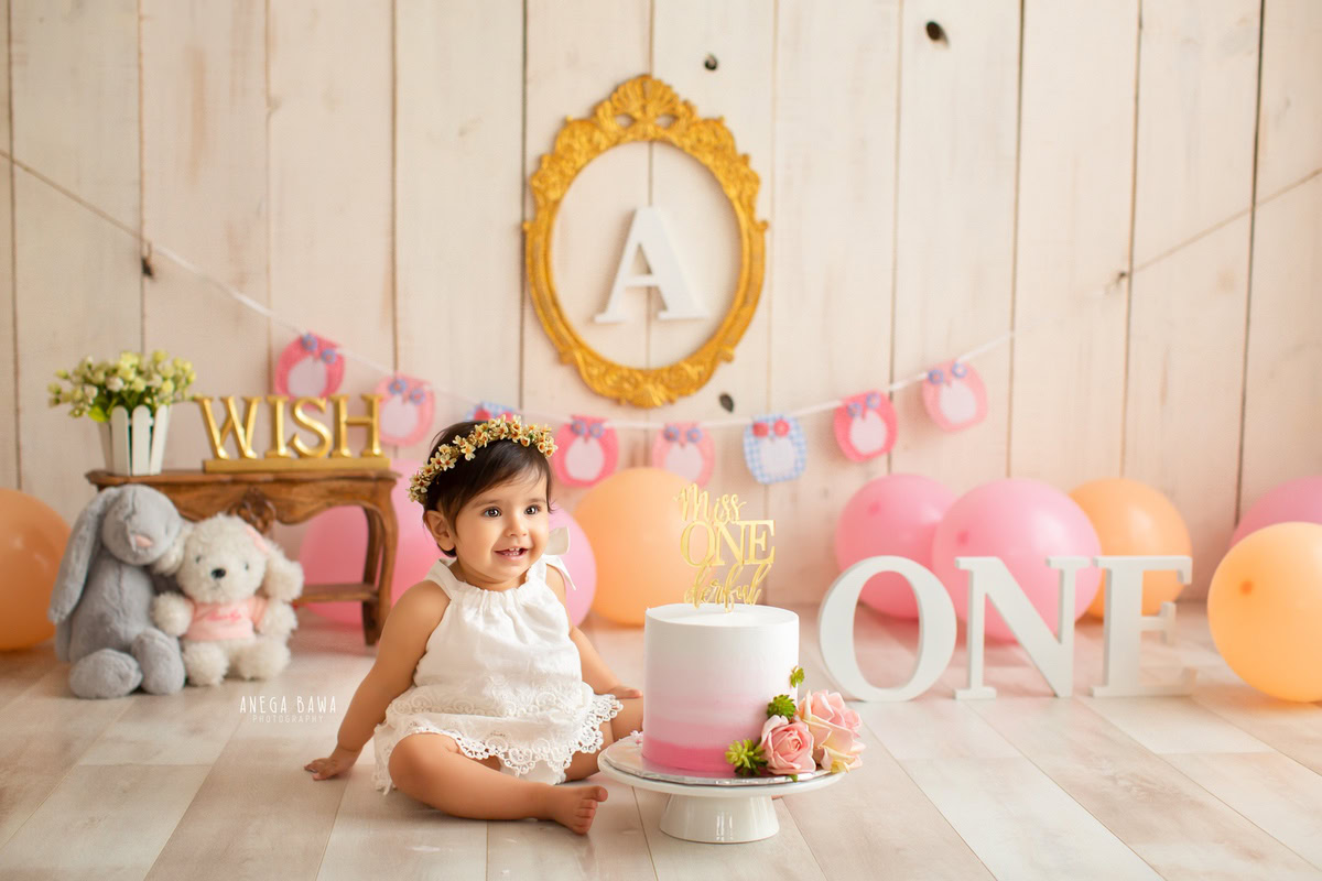 1 year old girl with a cake smash setup, featuring teddy bears, balloons, and pompoms against a beige backdrop. Celebrating her first birthday. Cake Smash photo shoot in Delhi, Gurgaon by Anega Bawa Family Photographer Gurgaon (Delhi NCR).