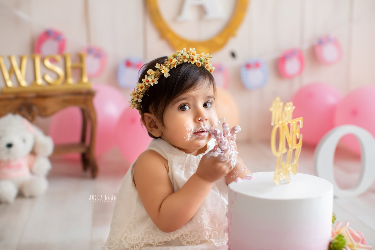 1 year old girl with cake smash wearing a tiara band surrounded by pink balloons and a beige backdrop. Captured during a lovely first birthday photography session in Delhi Gurgaon by the renowned family photographer Anega Bawa.