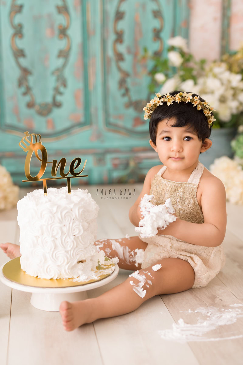 1 year old girl with a cake smash setup, featuring a tiara, green door backdrop, green vase, and white flowers. Celebrating her first birthday. Cake Smash photo shoot in Delhi, Gurgaon by Anega Bawa Family Photographer Gurgaon (Delhi NCR).