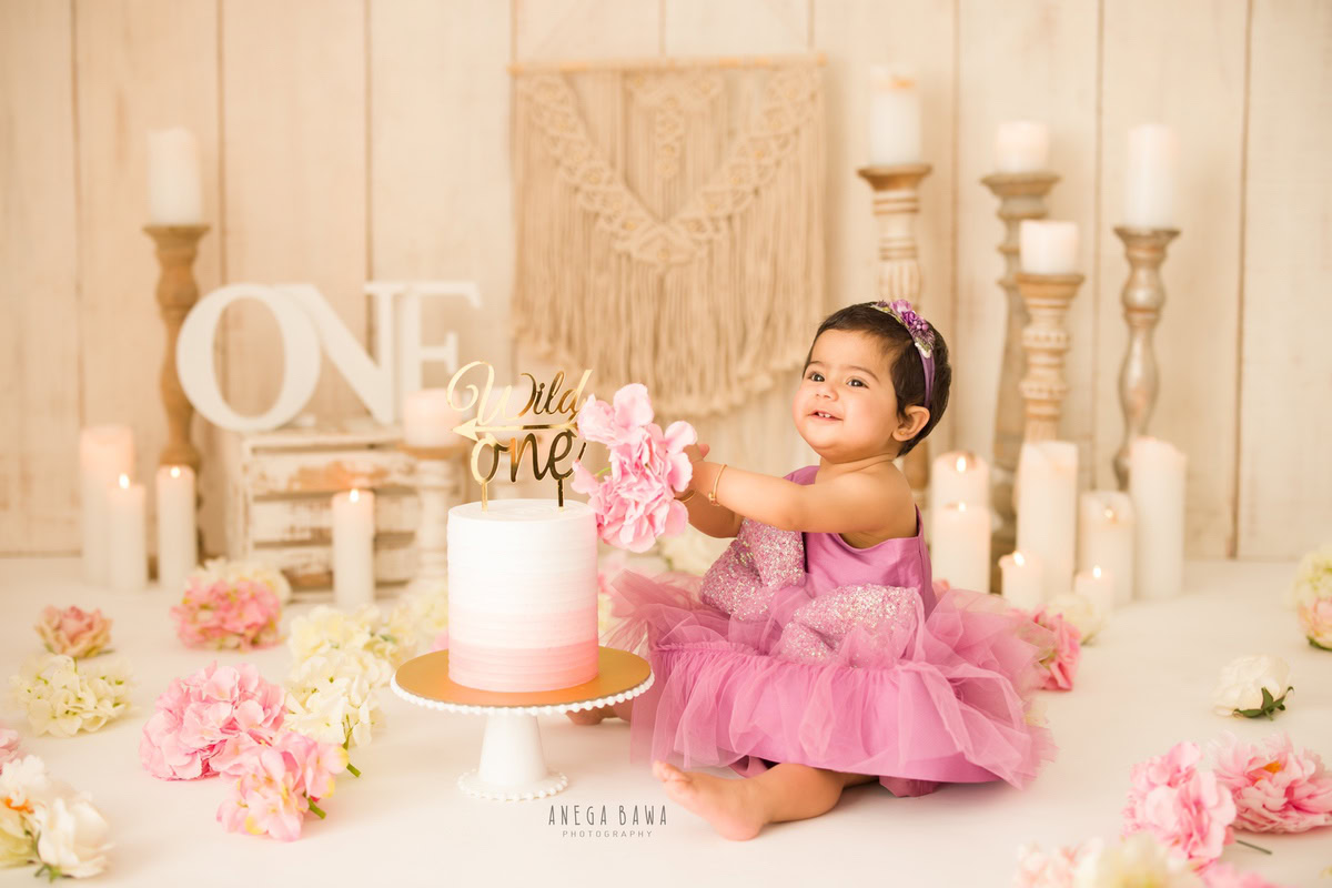 1 year old girl with a cake smash setup, featuring a candle stand, pink and ivory flowers, and a beige backdrop. Celebrating her first birthday. Cake Smash photo shoot in Delhi, Gurgaon by Anega Bawa Family Photographer Gurgaon (Delhi NCR).