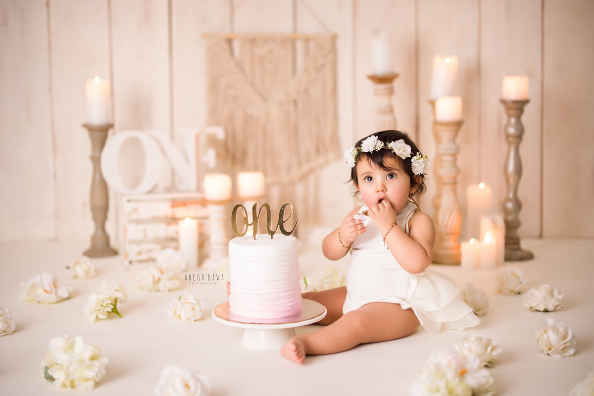 1year old girl with a cake smash setup, featuring a candle stand and white flowers, against a beige backdrop, captured by Anega Bawa during a Cake Smash photoshoot in Delhi, Gurgaon