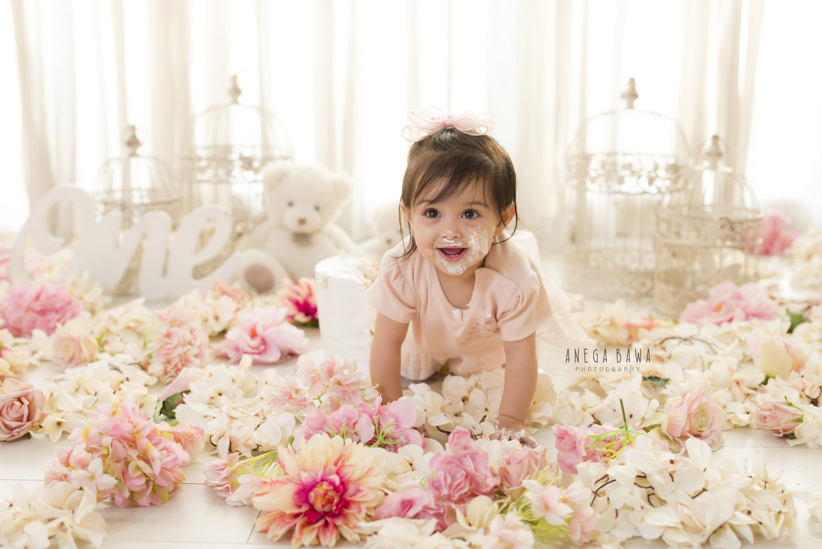 1year old girl crawling with cake on her face, surrounded by flowers on the floor and teddy bears, against a white backdrop, captured by Anega Bawa during a Cake Smash photoshoot in Delhi, Gurgaon
