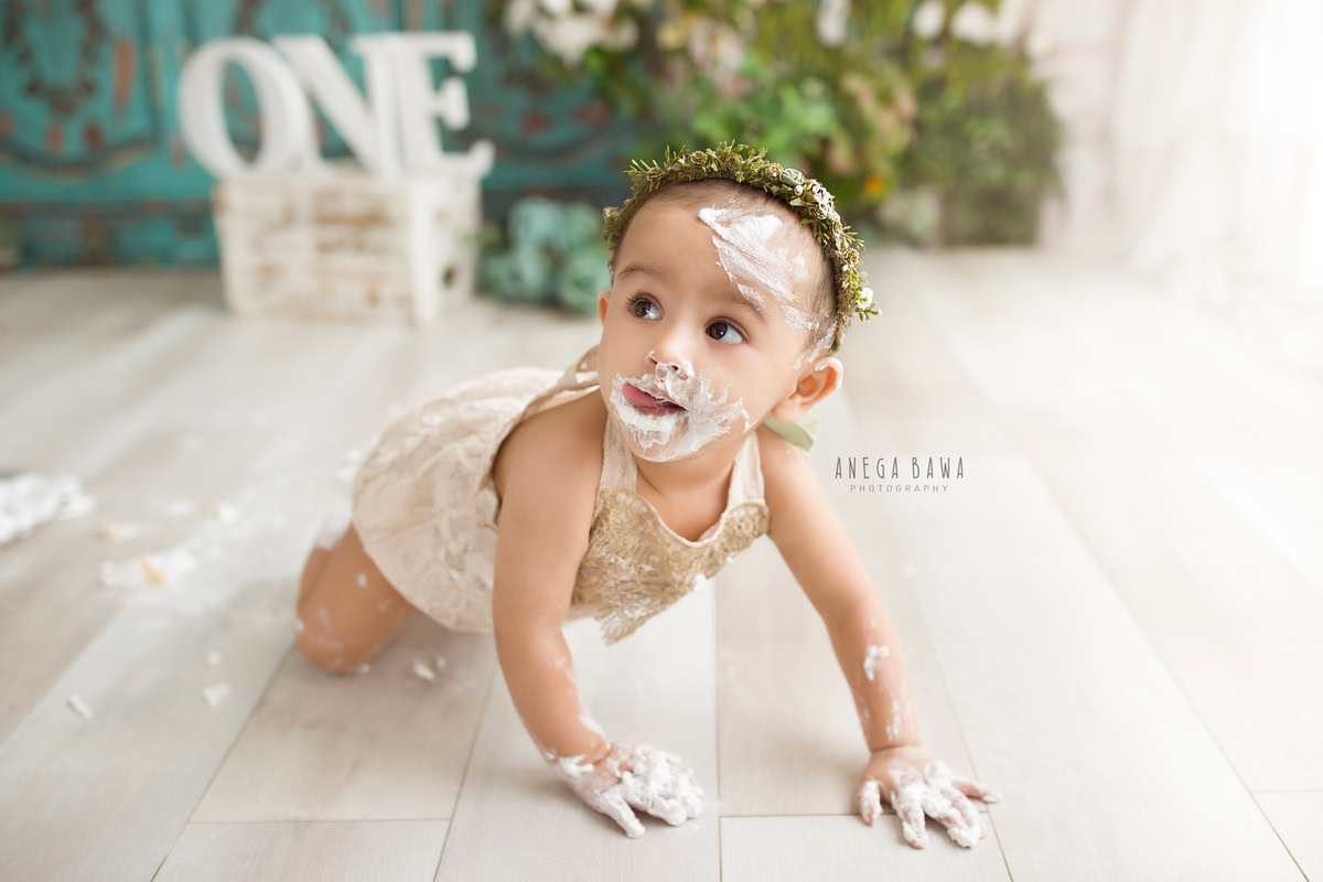 1 year old girl crawling with cake smash on her face against a green backdrop. Captured during a delightful first birthday photoshoot in Delhi Gurgaon by the renowned family photographer Anega Bawa.