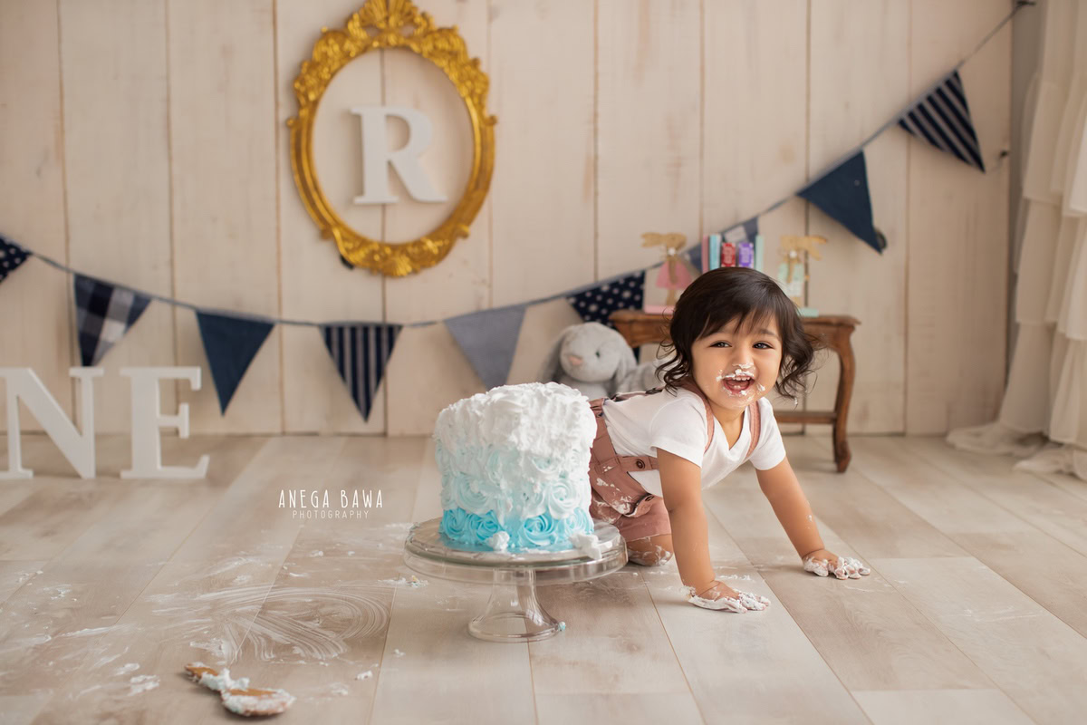 1 year old girl crawling on the floor with cake smash golden alphabet frame on the wall and a beige backdrop. Captured during a charming first birthday photography session in Delhi Gurgaon by the renowned family photographer Anega Bawa.
