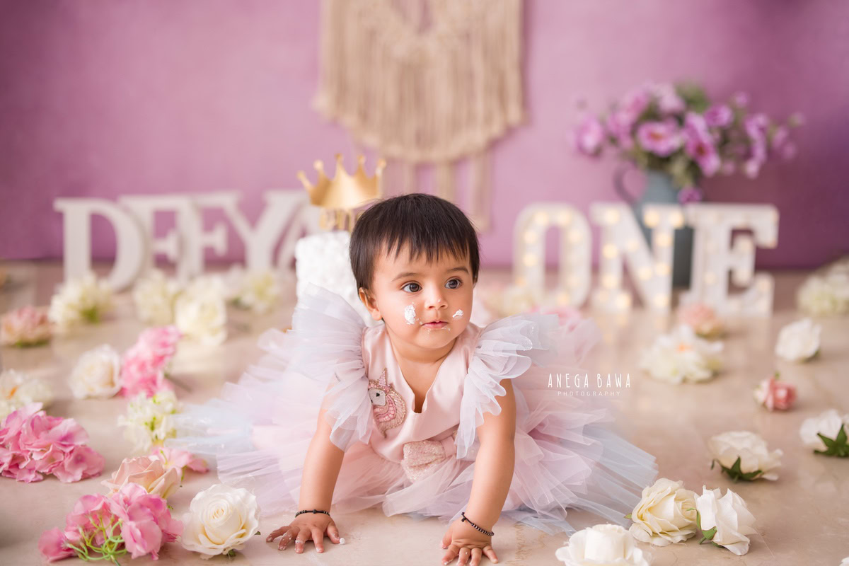 1year old girl crawling on the floor with pink and white flowers and a name frame, against a lavender backdrop with a cake smash setup, captured by Anega Bawa during a Cake Smash photoshoot in Delhi, Gurgaon