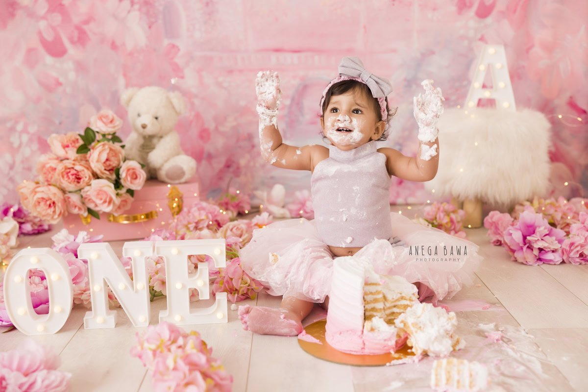 1 year old girl with cake smash on her face surrounded by pink and peach flowers teddybear and a pink backdrop. Captured during a delightful first birthday photo shoot in Delhi Gurgaon by the renowned family photographer Anega Bawa.