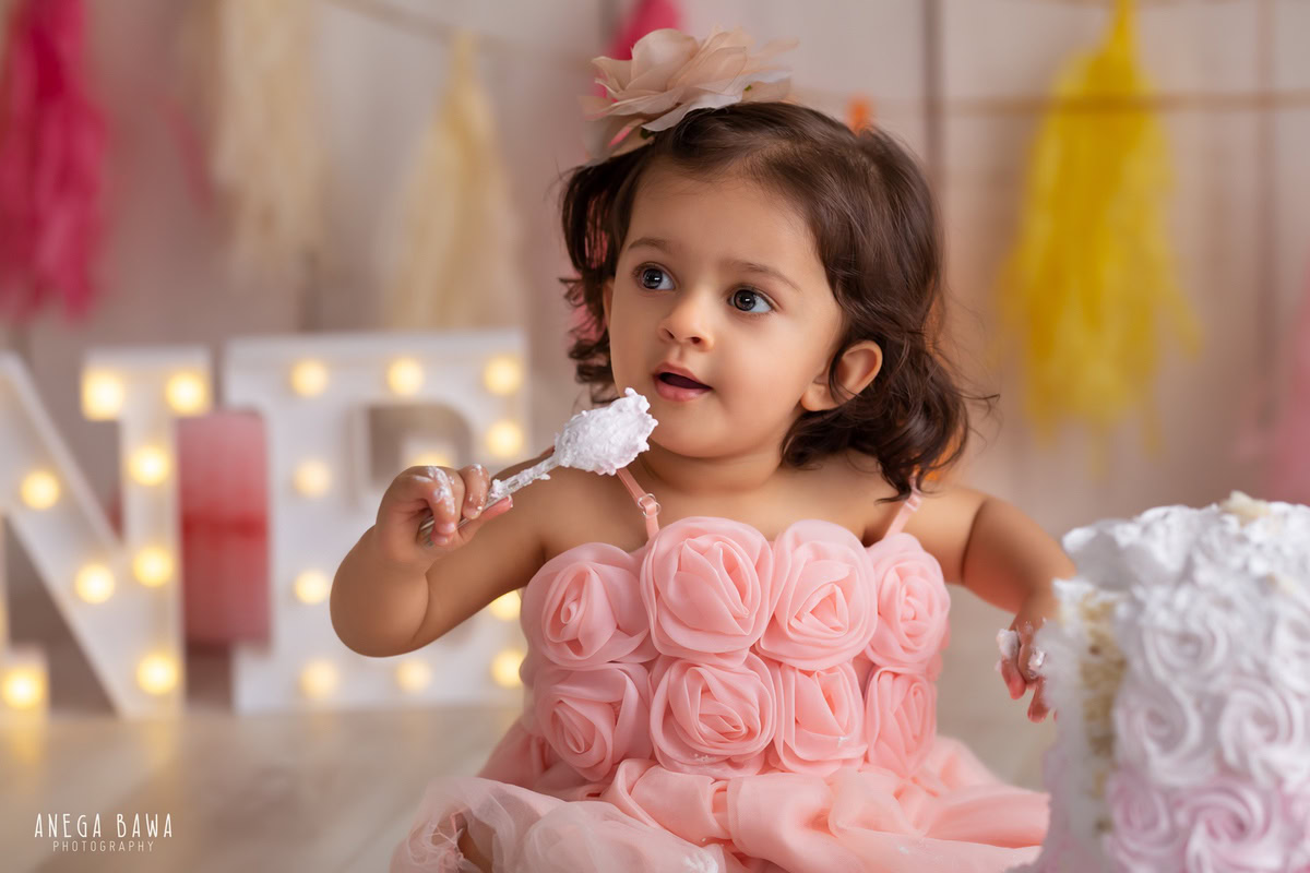 1 year old girl enjoying a cake smash setup, with a beige backdrop and multicolored fringes on the wall. Celebrating her first birthday. Cake Smash photo shoot in Delhi, Gurgaon by Anega Bawa Family Photographer Gurgaon (Delhi NCR).
