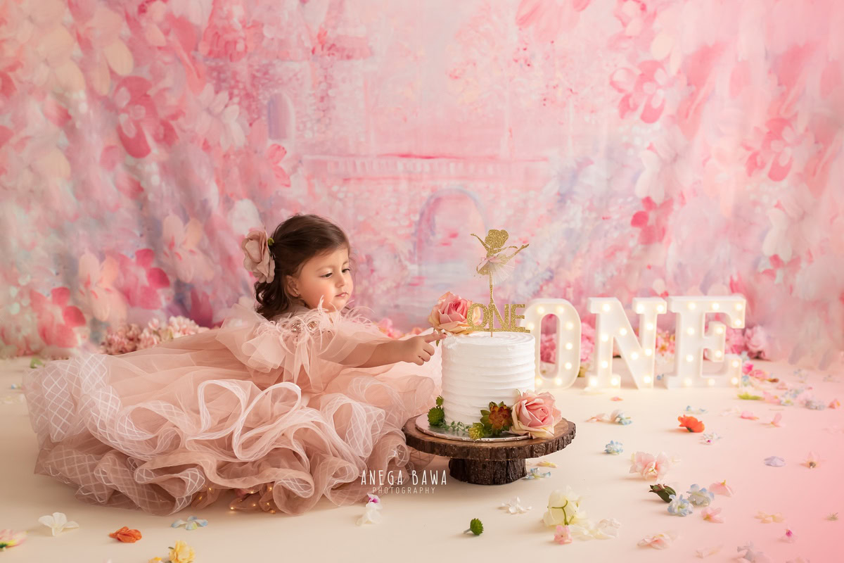 1 year old girl in a fairy dress with a cake smash setup, featuring a pink backdrop and flowers on the floor. Celebrating her first birthday. Cake Smash photo shoot in Delhi, Gurgaon by Anega Bawa Family Photographer Gurgaon (Delhi NCR).