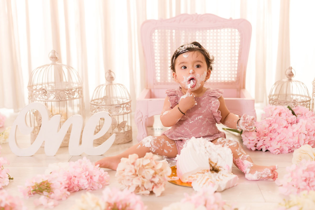1year old girl with flowers, castles, and a wooden pink chair against a white backdrop, featuring a cake smash setup, captured by Anega Bawa during a Cake Smash photoshoot in Delhi, Gurgaon